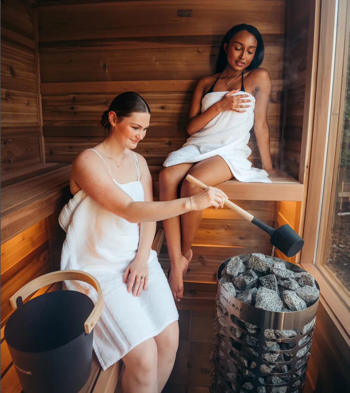 Two people on a cedar bench beside a stone-filled Homecraft sauna heater as one pours water