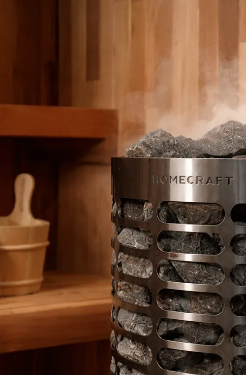 Sauna guides and resources Stone-filled Homecraft sauna heater steaming in a cedar room, with a wooden bucket and ladle on the bench