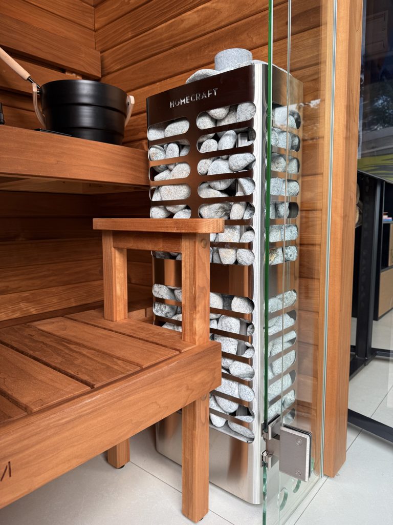 Homecraft Revive Slim sauna heater beside glass in a cedar room with a bench, bucket and pale stones