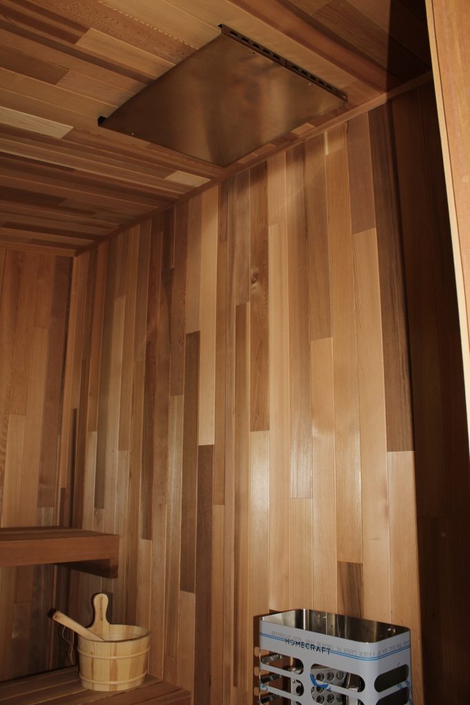 Cedar sauna interior with a ceiling heat shield, a bucket and ladle on the bench and a heater behind