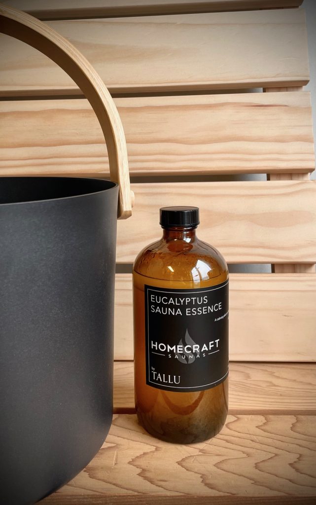 Amber eucalyptus sauna essence bottle on a cedar bench beside a black bucket with a wooden handle