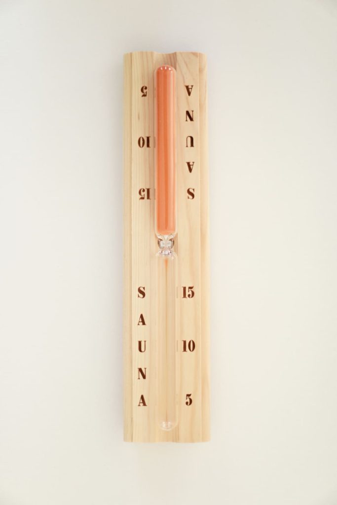 Wooden sauna sand timer with orange sand, minute markings and SAUNA text on a white wall