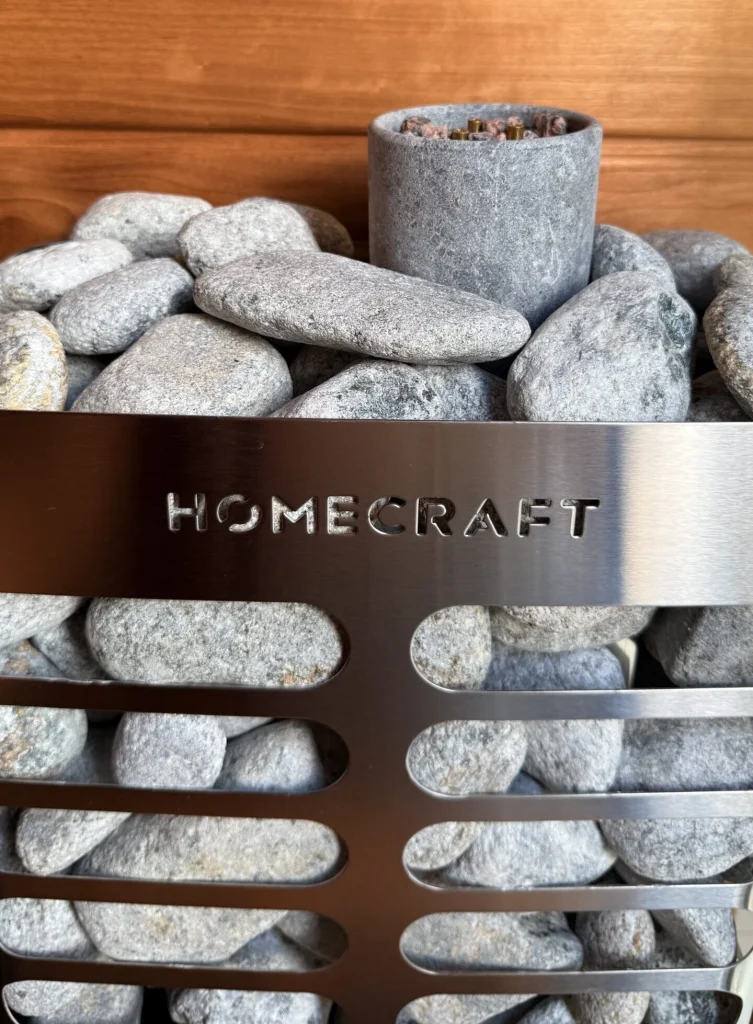Grey sauna stones in a steel heater cage with a soapstone cup and cedar wall behind