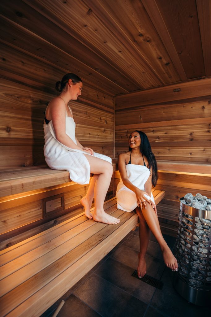 Two people in a cedar sauna Two women seated in a Canadian cedar sauna kit with tiered benches, under-bench lighting and a stone heater