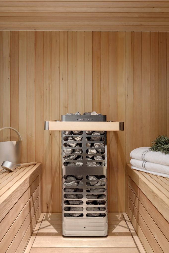 Homecraft Revive sauna heater with steel guard and wooden rail beside towels, watering can and plant