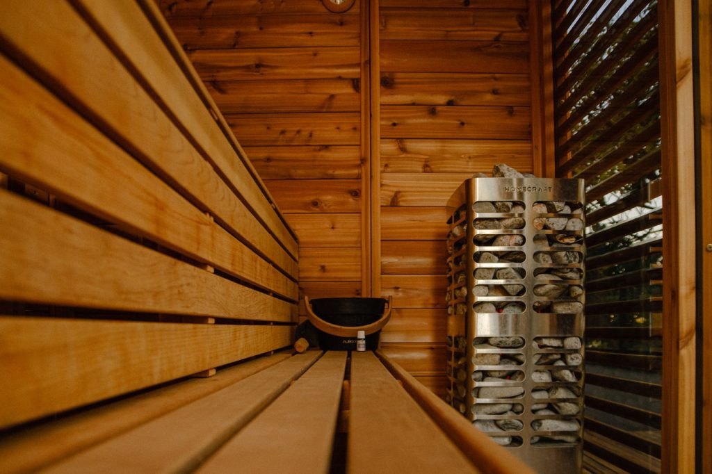 Cedar sauna interior and heater Canadian cedar sauna kit interior with bench, wooden bucket, tall steel heater and dark slatted wall panel