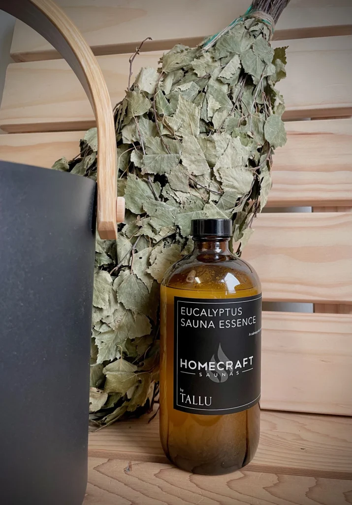 Amber eucalyptus sauna essence bottle on a cedar bench beside a dried birch whisk and black bucket