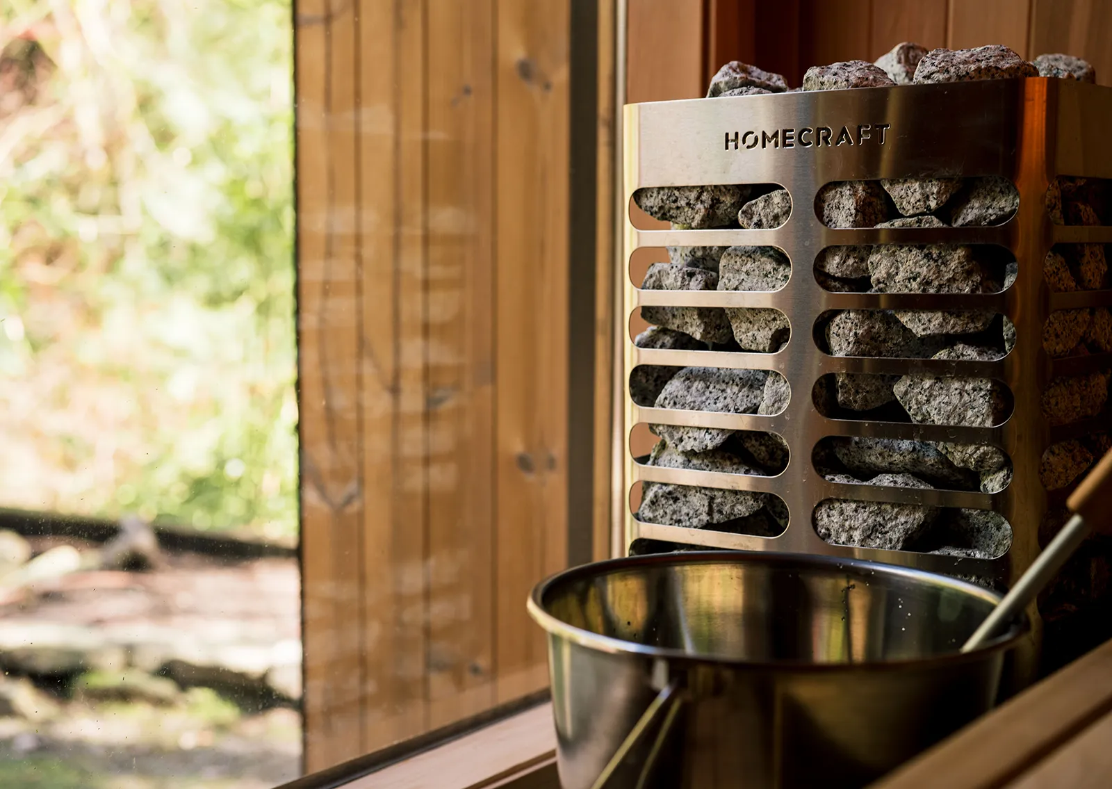 Homecraft sauna heater packed with stones beside a stainless bucket and ladle, with a window behind