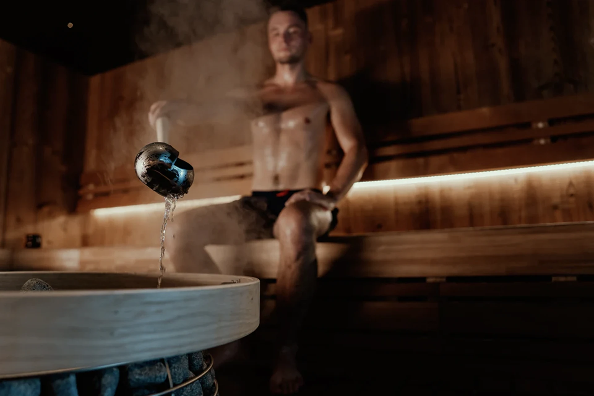 Man seated on a cedar sauna bench as water pours into a wooden tub under dim bench lighting
