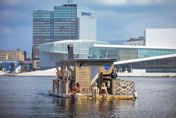 Bathers relaxing on a floating sauna in a city harbour, with an opera house and glass towers behind