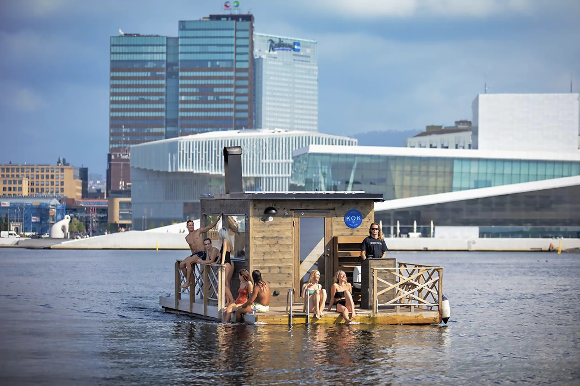 Bathers relaxing on a floating sauna in a city harbour, with an opera house and glass towers behind