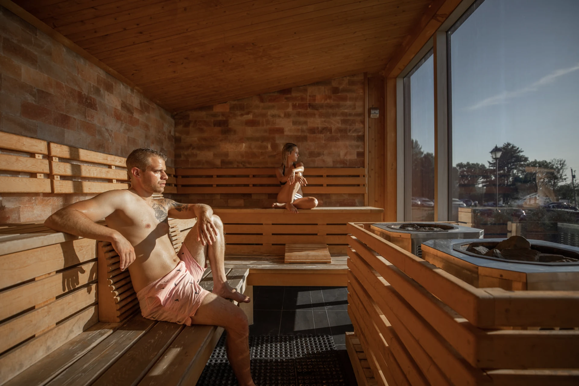 Man seated on a cedar sauna bench beside a window overlooking a wood-fired hot tub among pines
